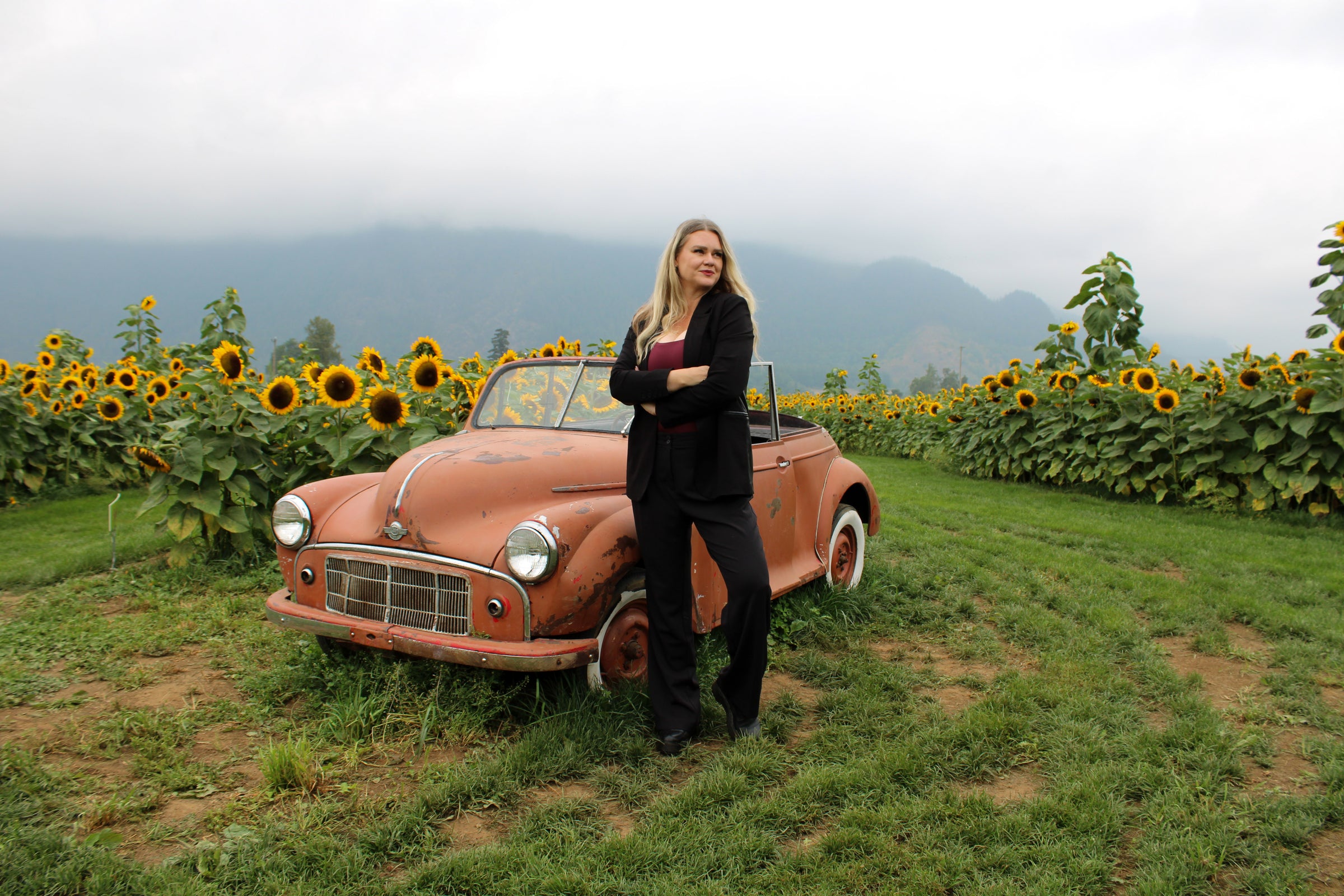 Leanne standing in a sunflower field in a black tailored suit, leaning on a vintage car.